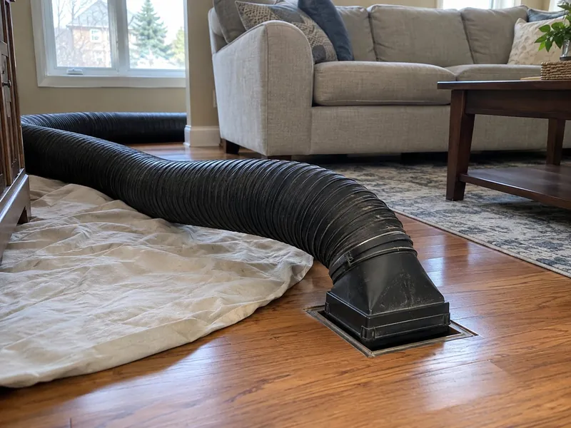 A thick black duct-cleaning vacuum hose is connected with a square adapter to a floor register in a living room, beside a drop cloth on the hardwood floor, with a sofa behind.